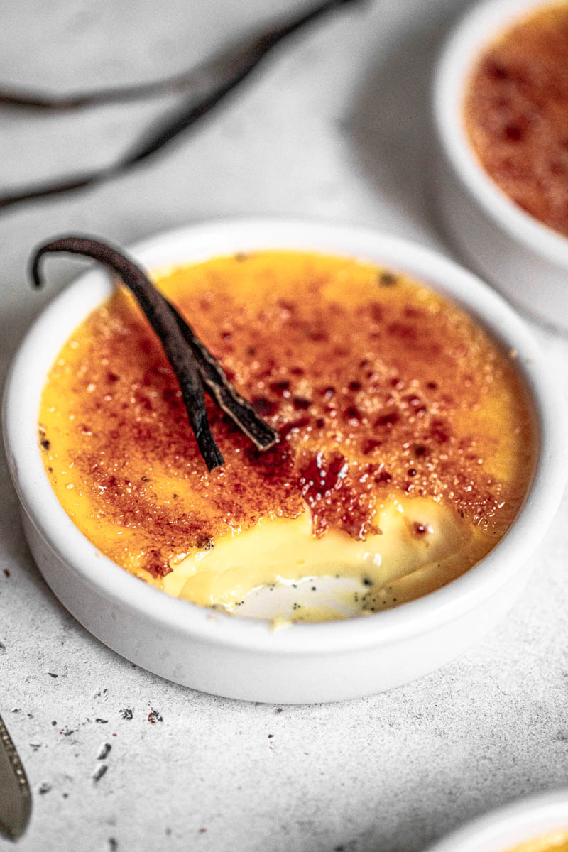 Vanilla bean creme brûlée, baked and torched, in an round ramekin, with another crème brûlée behind it and a dried vanilla bean on top.