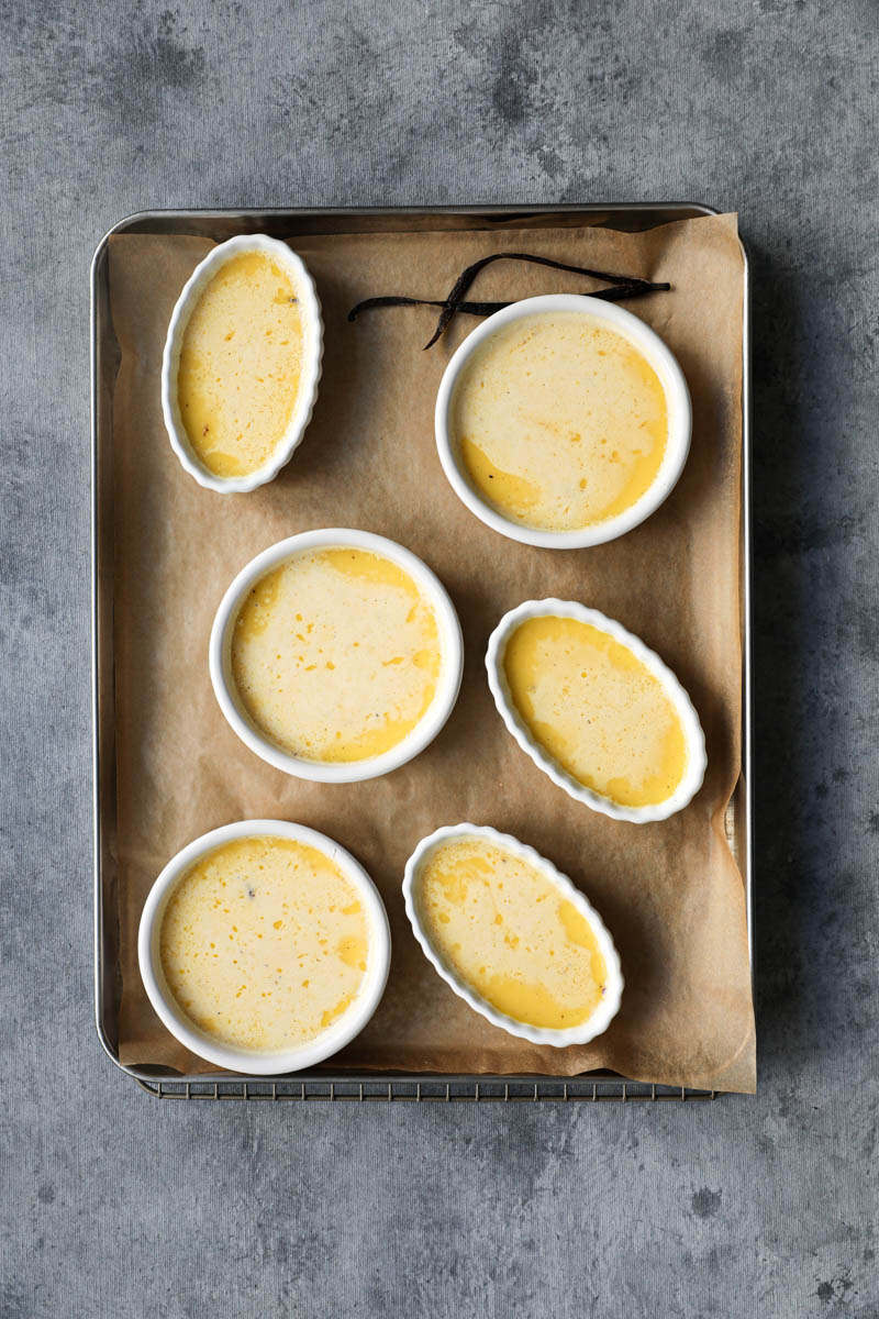 In the process of making vanilla bean crème brûlée: the baked and chilled crème brulee in white oval and round ramekins on a baking tray lined with parchment paper.