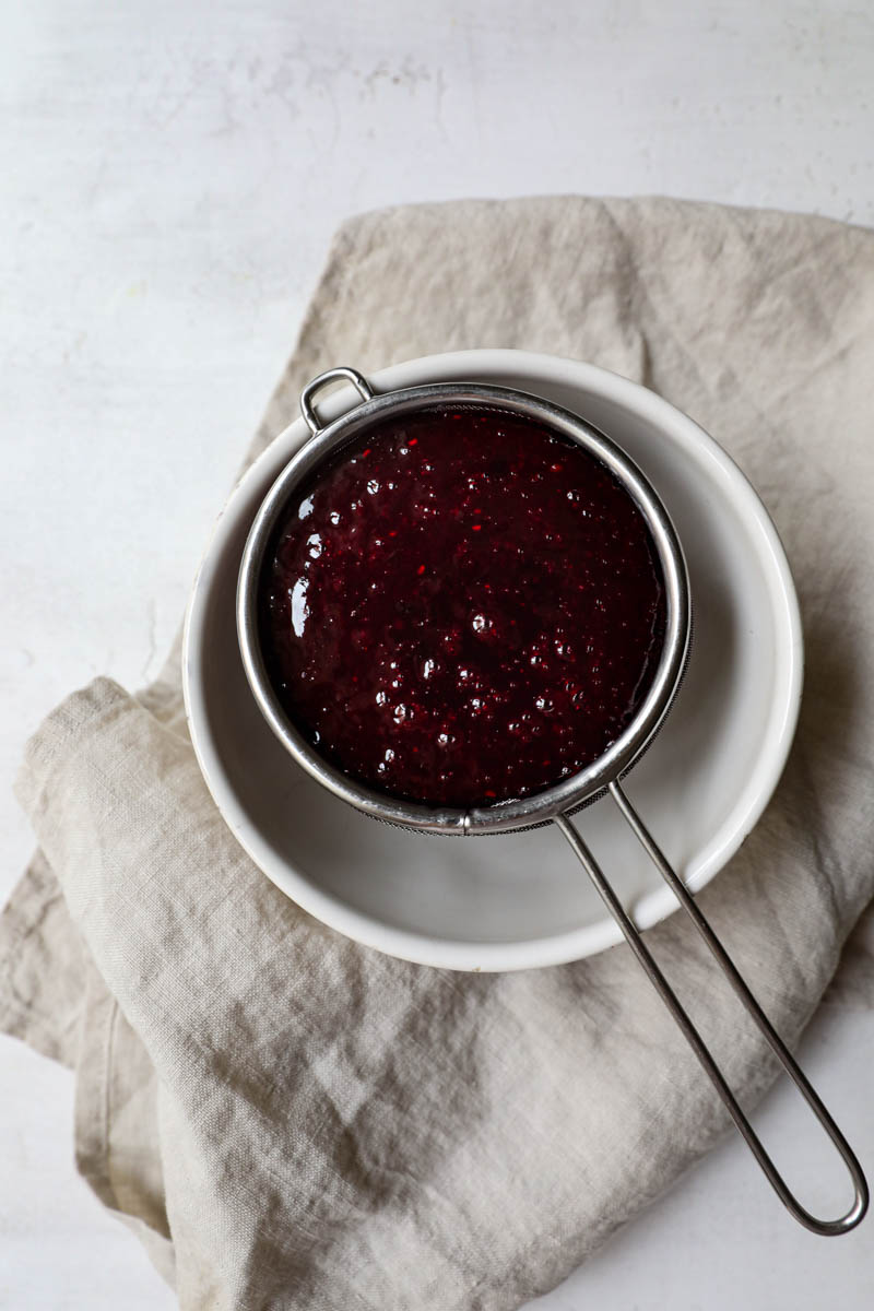 Berry sauce being strained over a small fine mesh strainer placed on top of a white bowl.