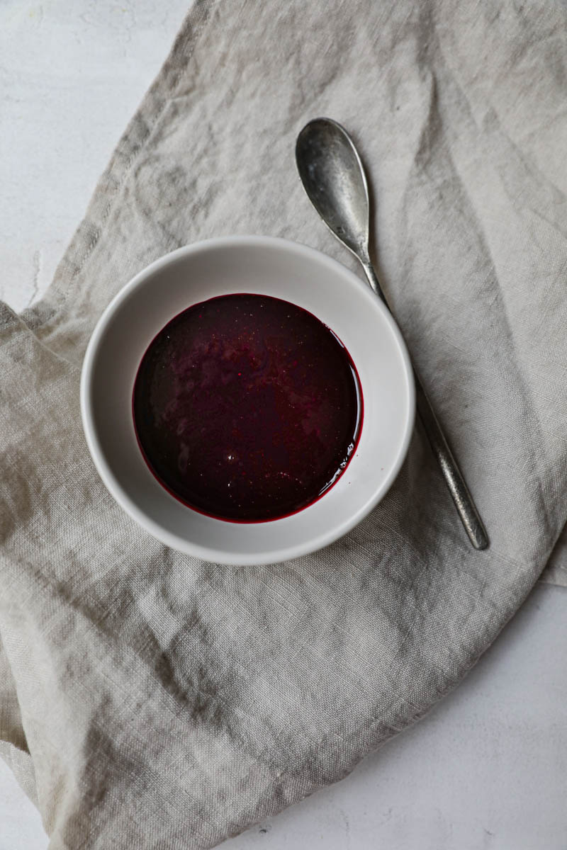 Berry sauce inside a small bowl on a beige linen cloth with a spoon on the side.
