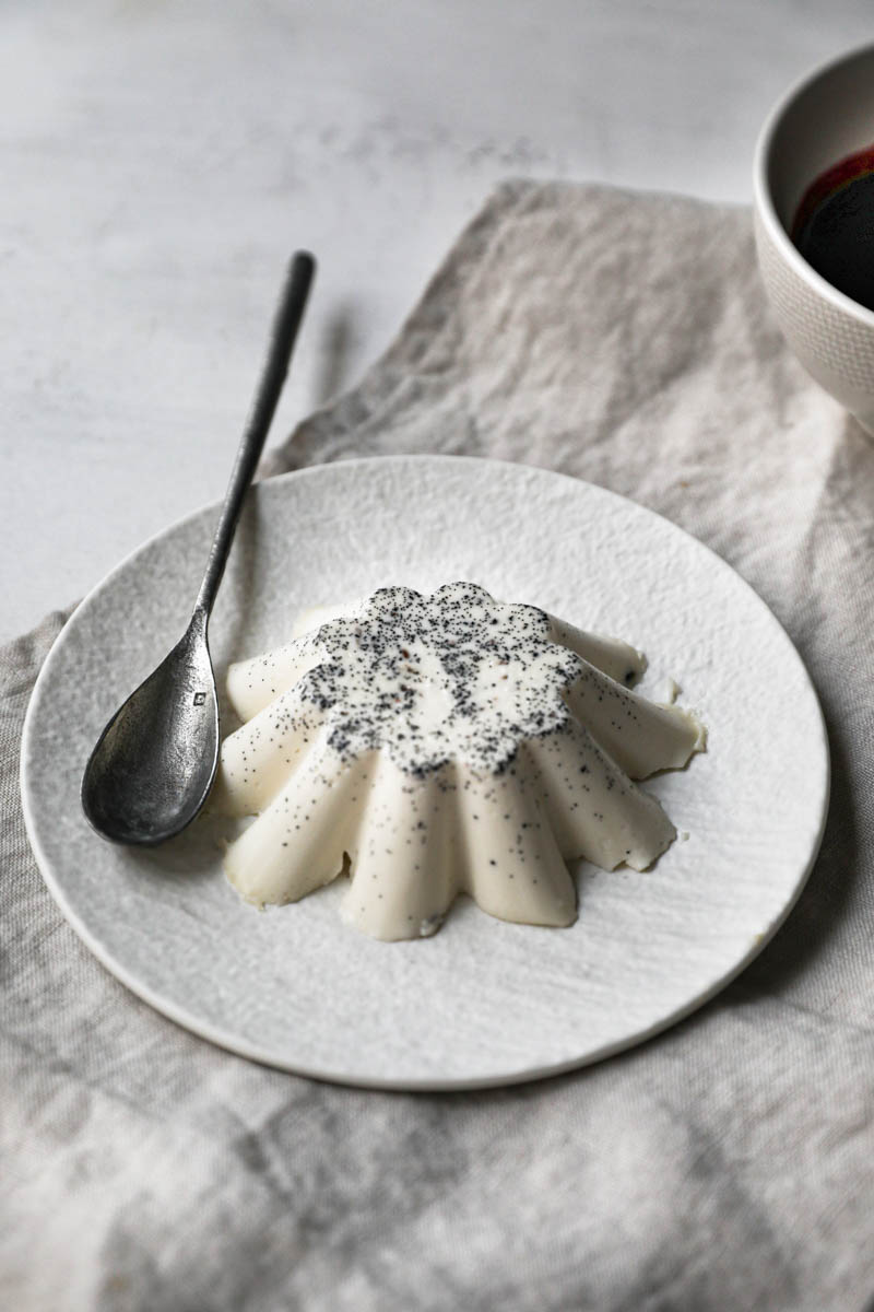 Panna cotta chilled and unmoulded on white plate on top of a beige linen cloth with a spoon on the side.