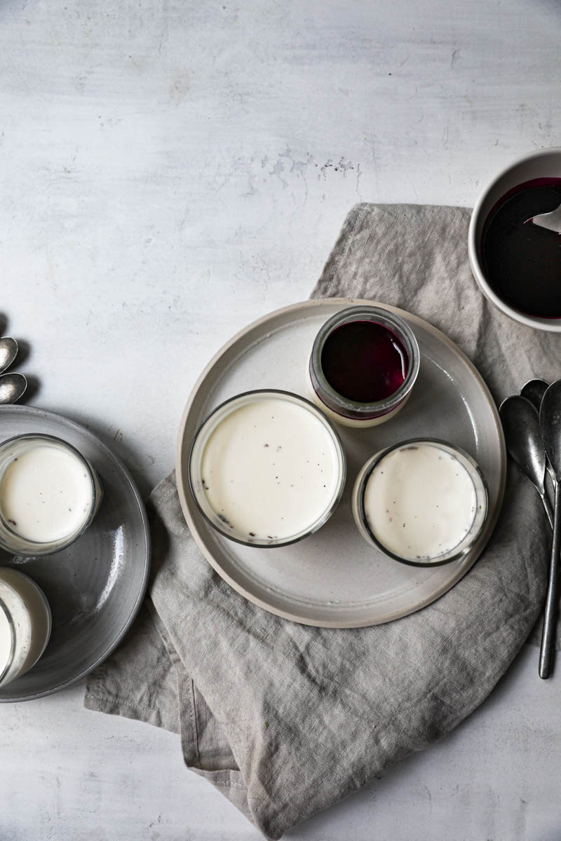 The glass containers, placed on 2 plates, filled with the panna cotta, chilled and some covered with the berry sauce, as seen from above. All is on top of a beige linen cloth.