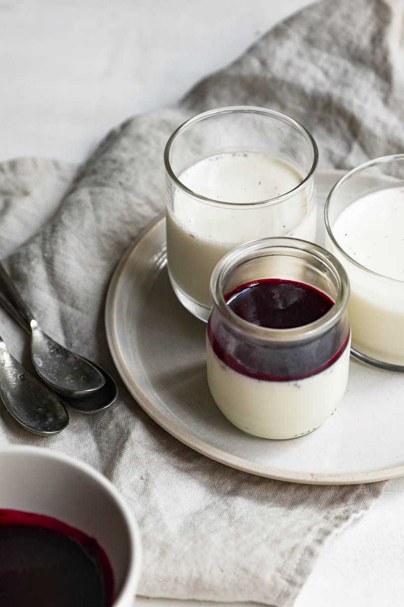 The glass containers, placed on 2 plates, filled with the panna cotta, chilled and some covered with the berry sauce, as seen from the front. All is on top of a beige linen cloth.