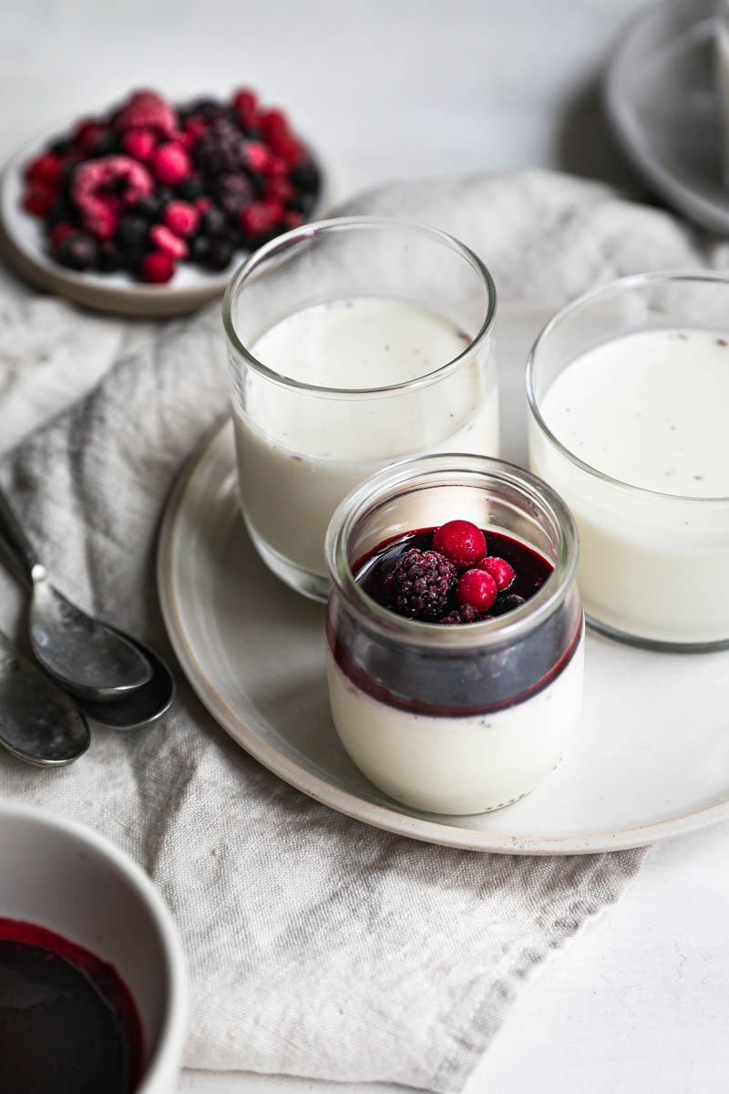 The glass containers, placed on 2 plates, filled with the panna cotta, chilled and some covered with the berry sauce and some berries, as seen from the front.