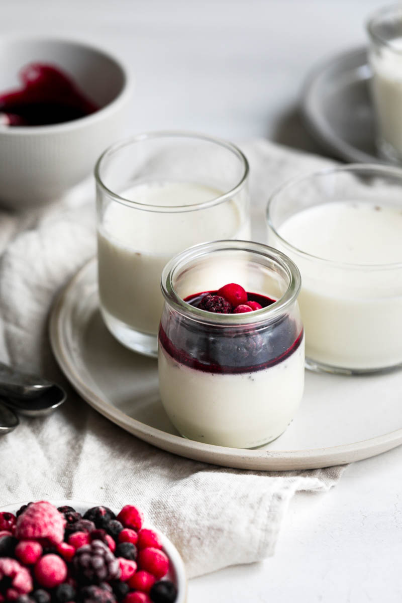 The glass containers, placed on 2 plates, filled with the panna cotta, chilled and some covered with the berry sauce and some berries, as seen from the front, placed on top of a beige linen.