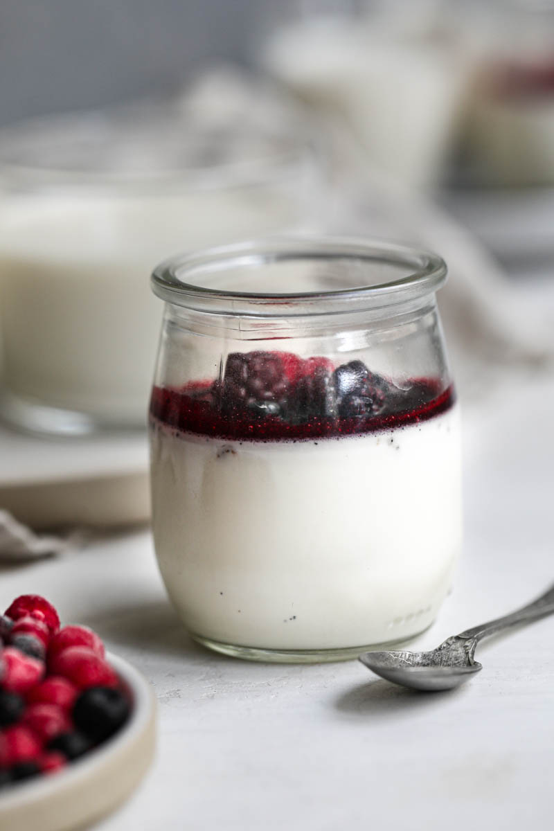 Closeup of one small glass filled with panna cotta, berry sauce and red berries with a spoon on the side and a plate with berries on the bottom left corner.