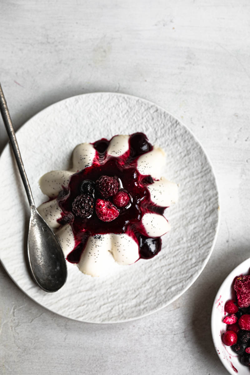 Panna cotta chilled and unmoulded on white plate with a spoon on the side, with a small plate with berries in the bottom right corner, as seen from the top.