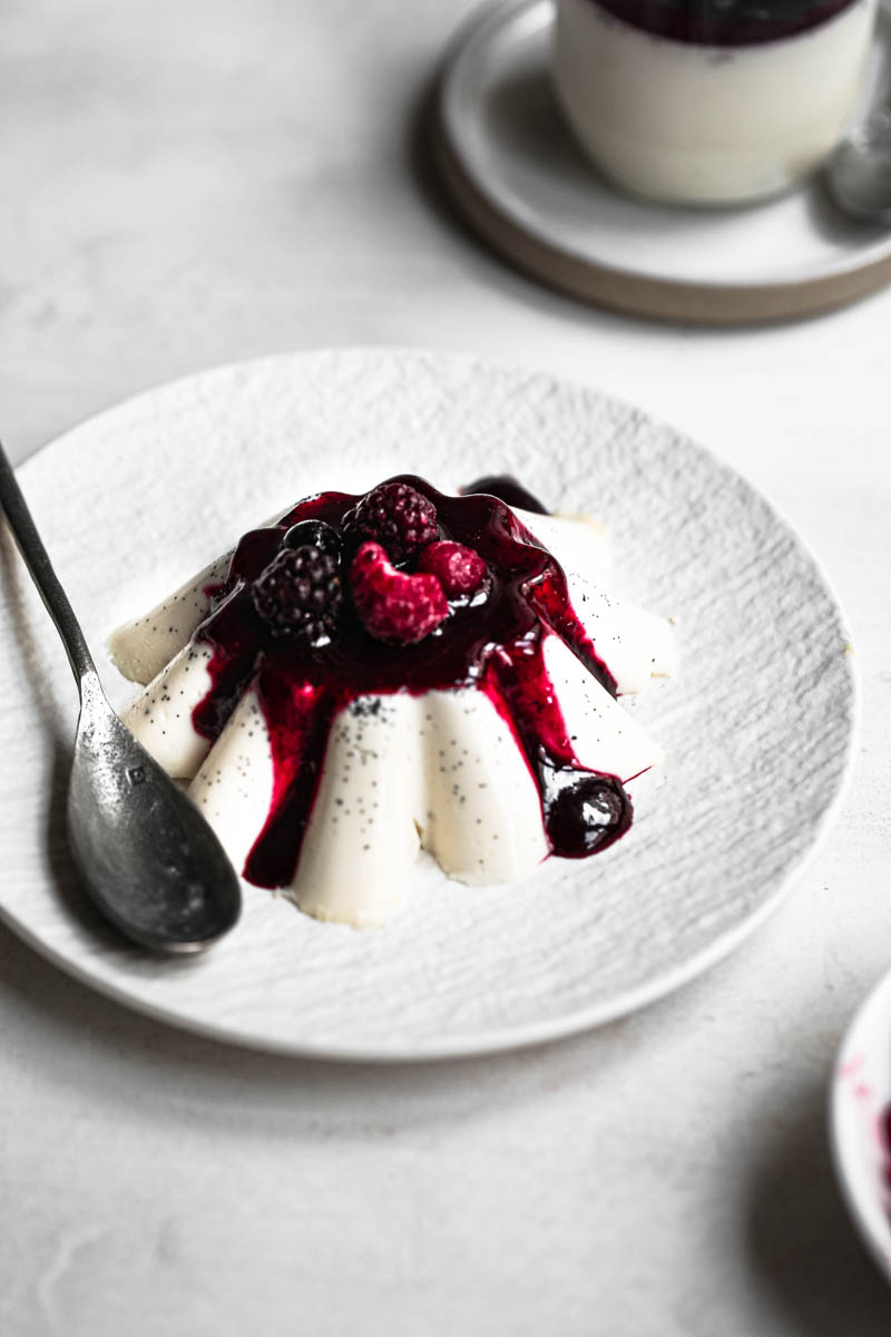 Unmoulded panna cotta in the shape of a flower over a white plate topped with berry sauce and fresh berries.