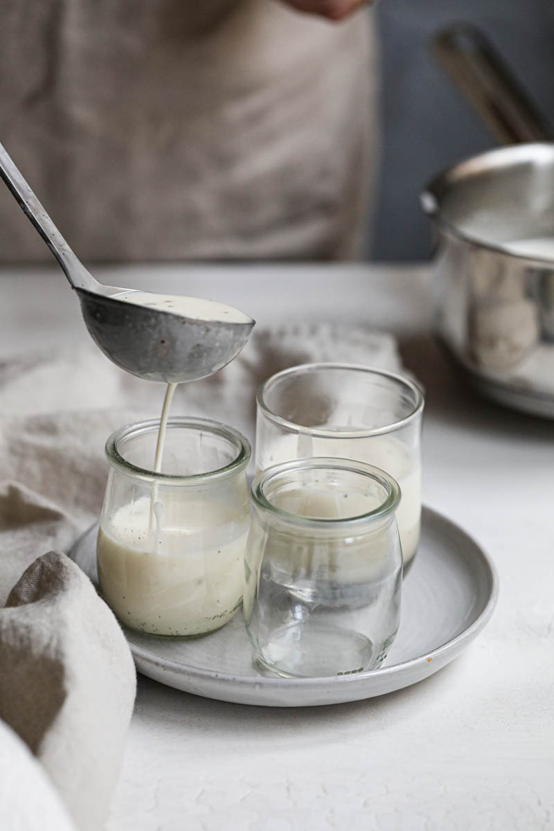 A hand filling small glass containers with vanilla panna cotta with a ladle.