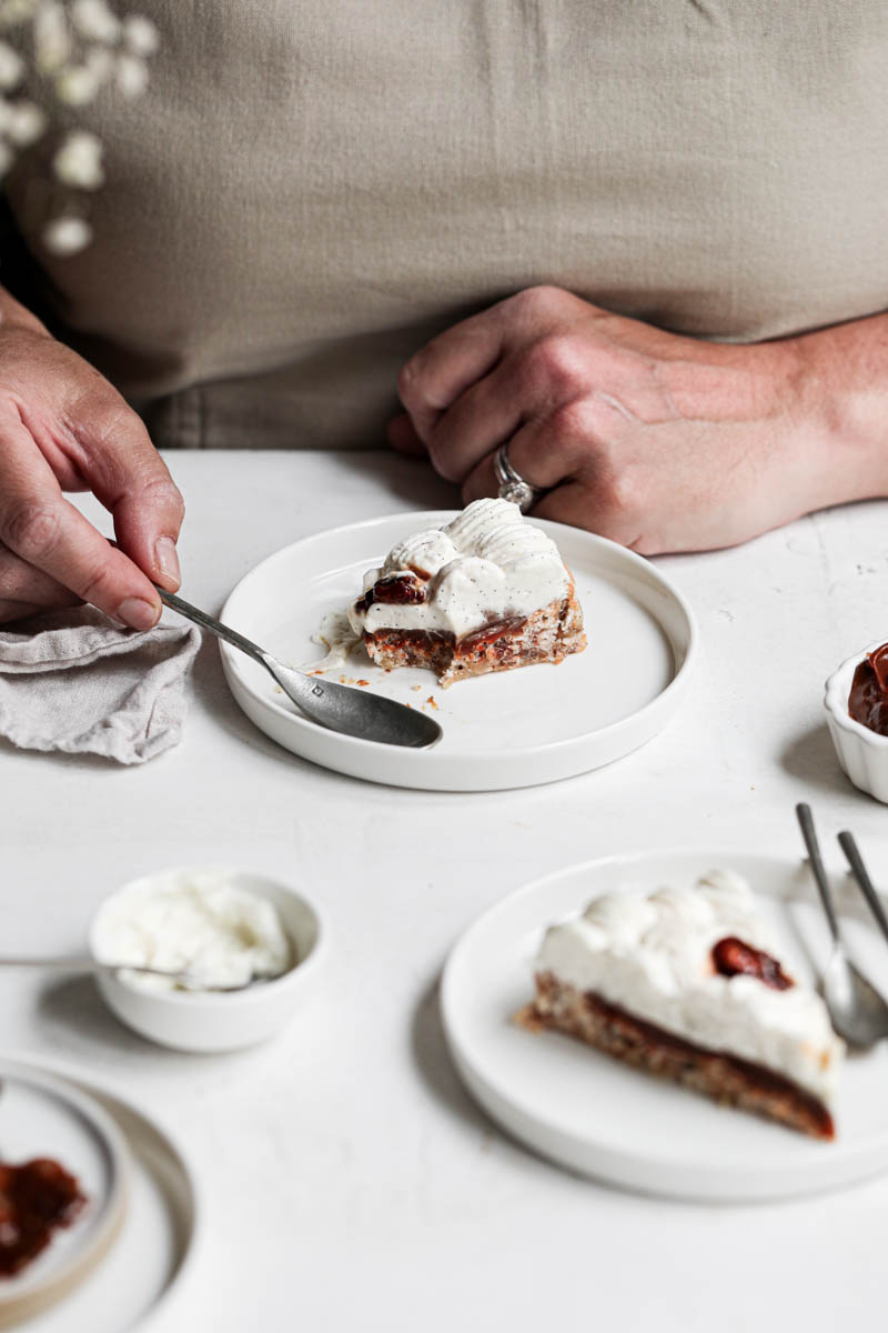 1 hand holding a spoon about to get a piece of the walnut dacquoise cake on plate in front of them, with another slice on a small plate on the side.