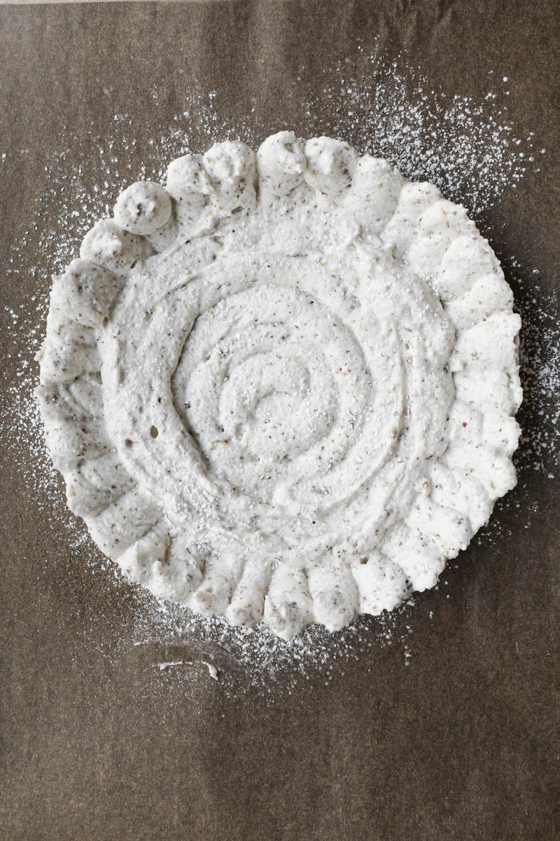 The dacquoise cake batter inside the cake ring placed on a baking sheet lined with brown parchment paper sprinkled with powdered sugar.