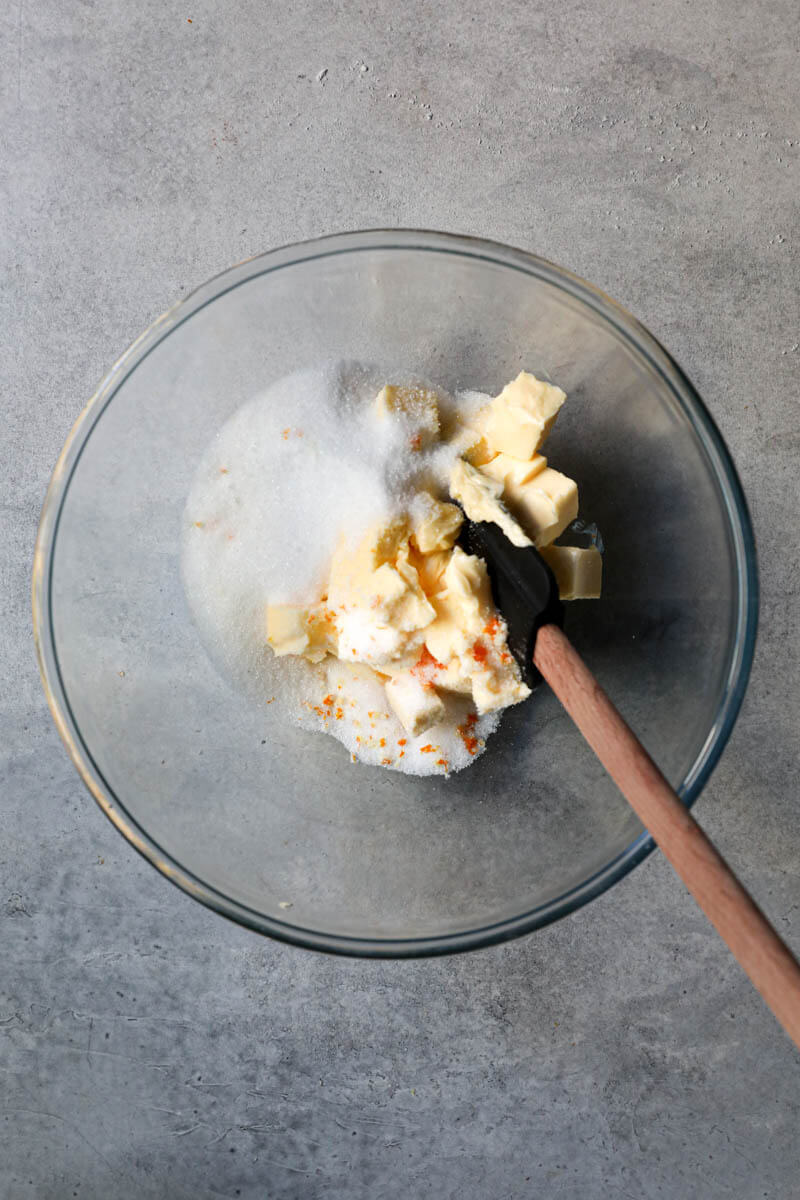 Softened butter, sugar and orange zests in a bowl with a black spatula
