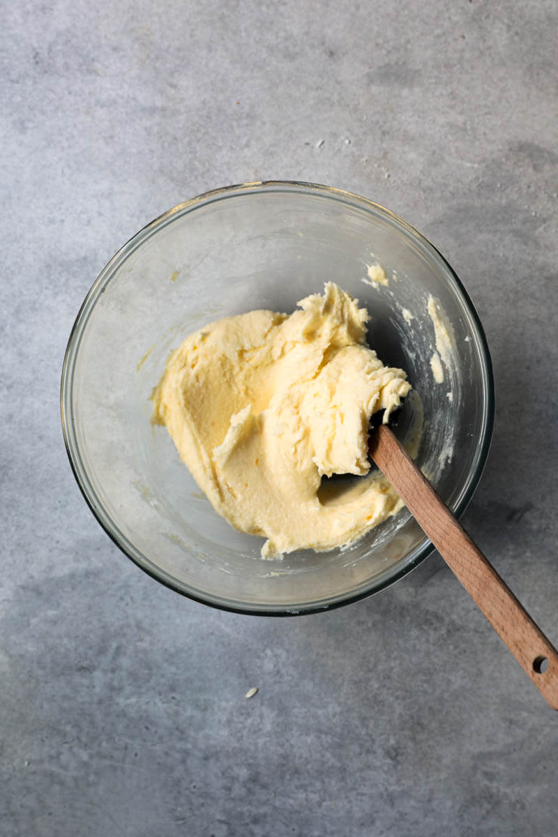 The softened butter, sugar and orange zests in a bowl with a black spatula after mixing