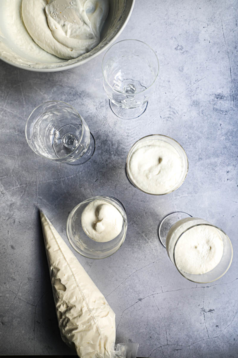 4 glasses filled with the white chocolate mousse and a pipping bag on the side and a bowl with the mousse on the top corner of the frame.