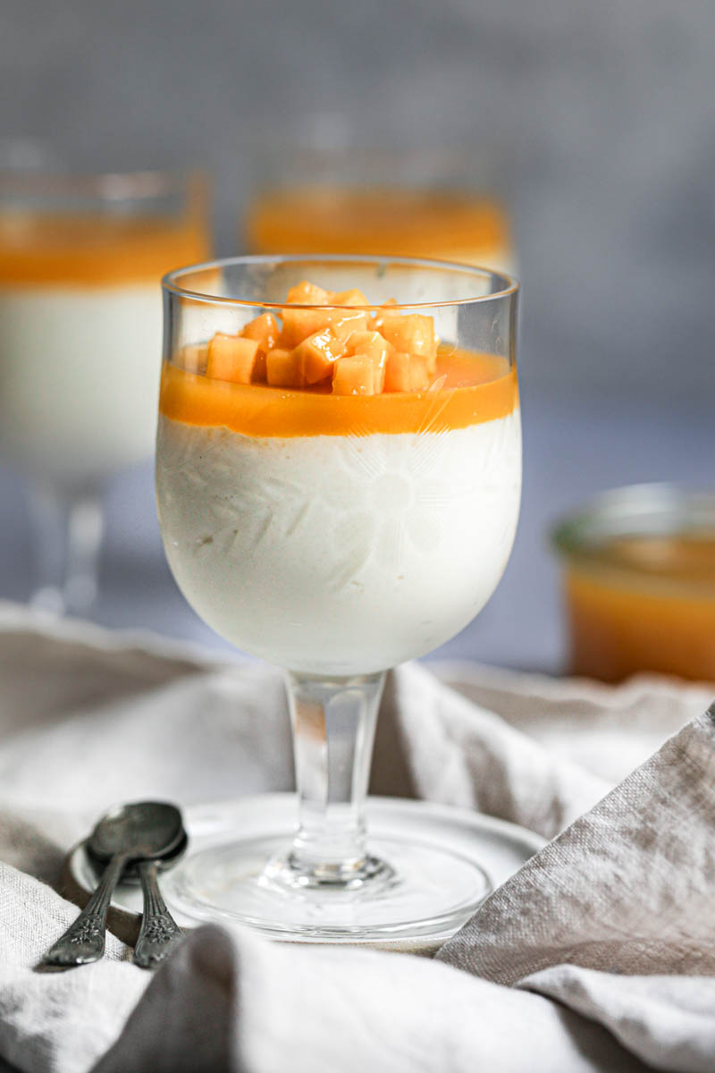 1 wine glass filled with white chocolate mousse and fresh mango and mango jelly on a small white plate with 2 spoons on the side and placed on a beige linen.