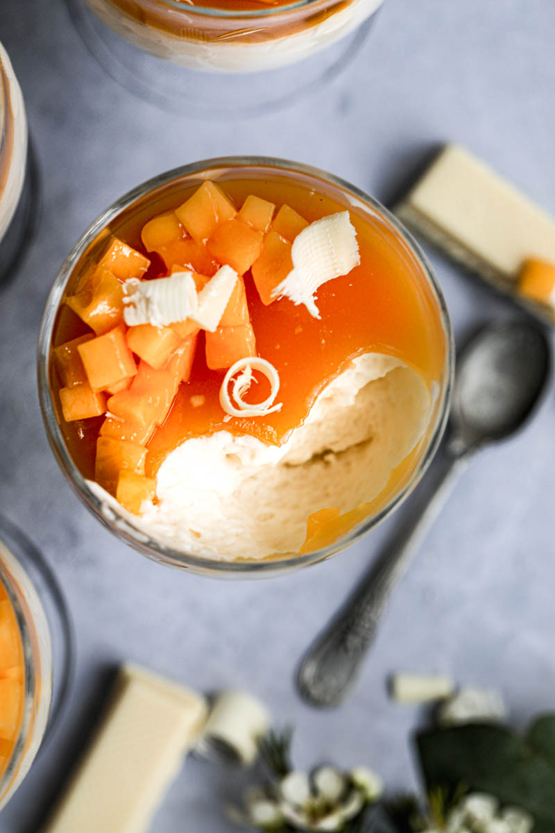 A closeup of 1 wine glass filled with white chocolate mousse and fresh mango and mango jelly as seen from the top, with one mousse missing a bite.