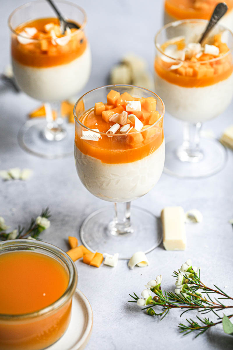 White chocolate mousse in a wine glass topped with mango jelly and fresh mango, with other glasses in the back and white chocolate and some leaves on the surface.