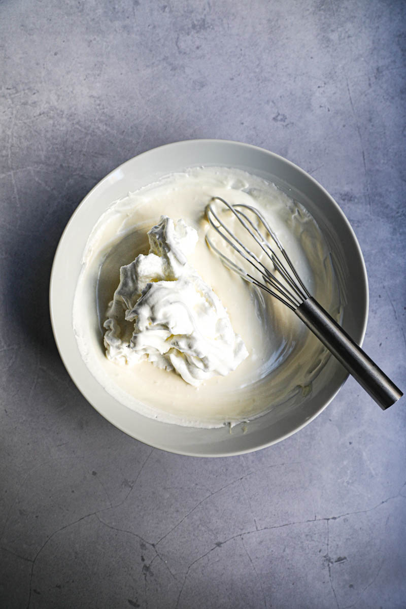 White chocolate ganache and whipped cream mixed together with one third of the beaten egg whites waiting to be mixed in and a whisk inside the bowl.