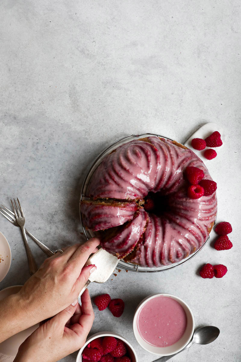 White chocolate raspberry cake with a pink glaze as seen from above with 2 hands pulling a slice from the side.