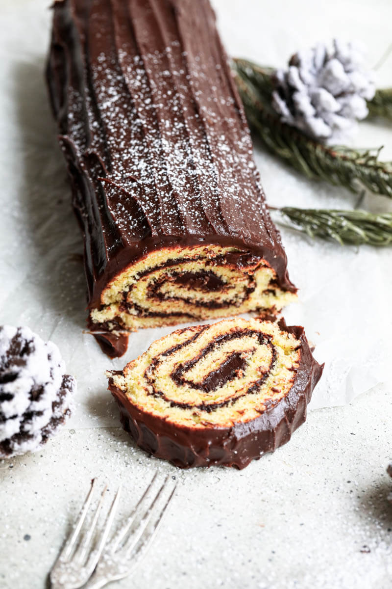 Sliced yule log cake with 2 forks on the left bottom corner and some pinecones to the right