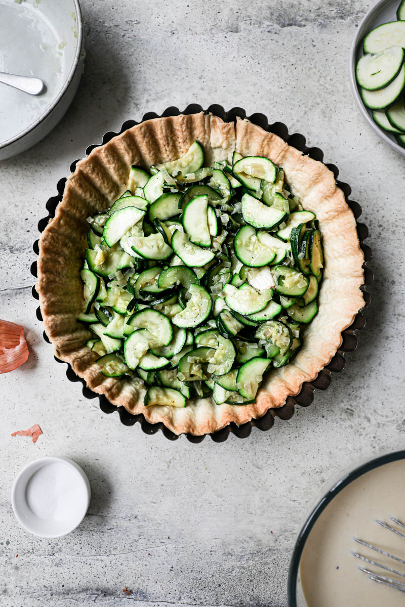 The blind baked quiche crust filled with the cooked zucchini and shallots with some plates and bowls on the side.