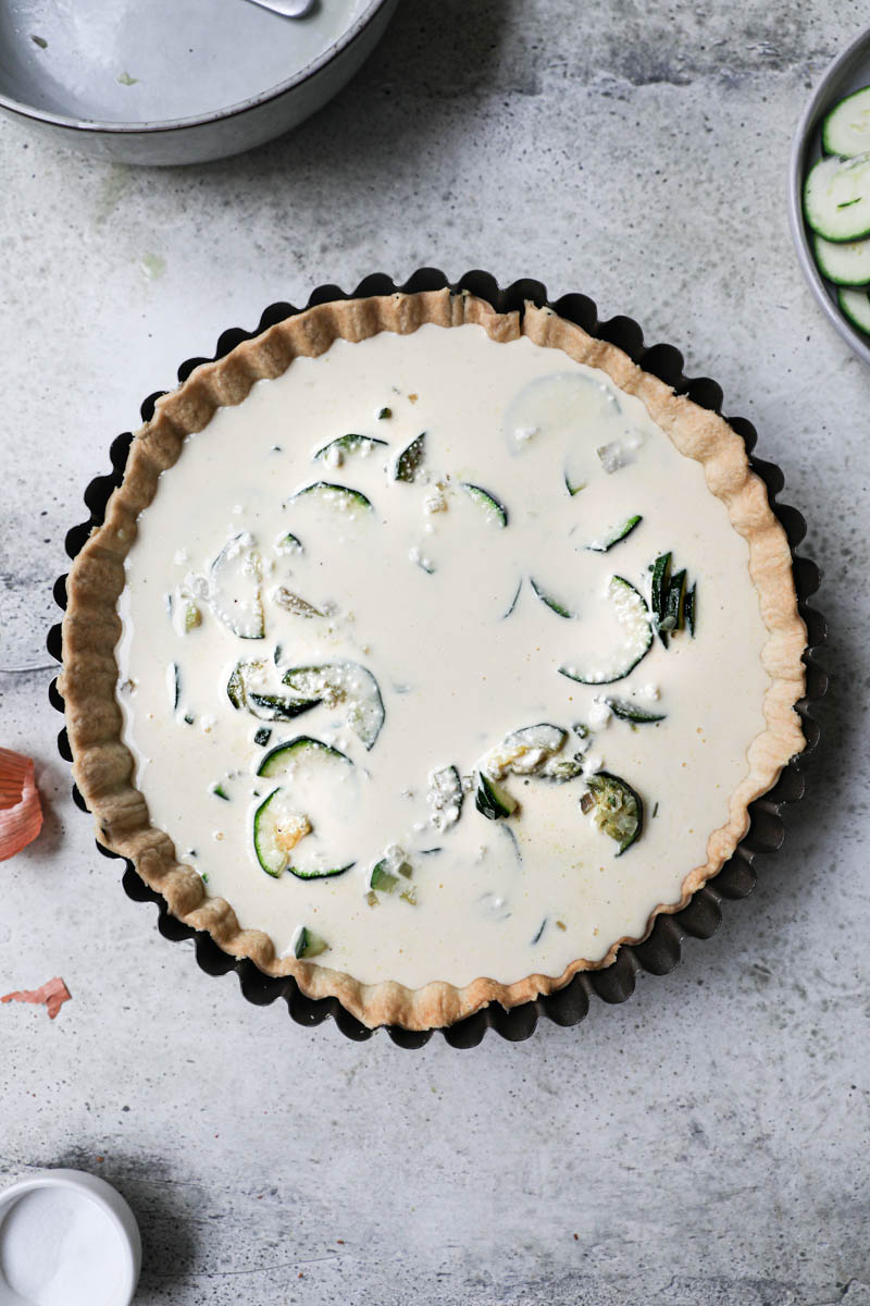 The blind baked quiche crust filled with the cooked zucchini and shallots covered with the quiche batter with some plates and bowls on the side.