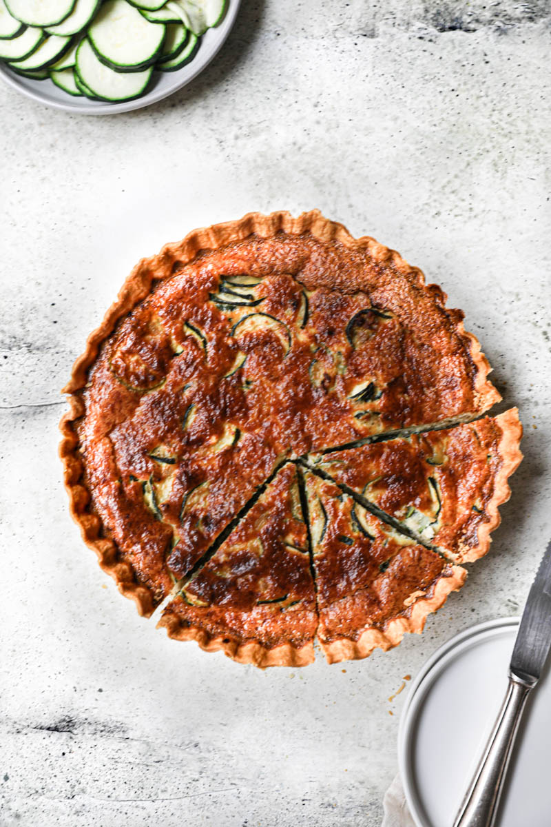 The whole zucchini quiche sliced with a knife on the side and plate with sliced zucchini in the top left corner.