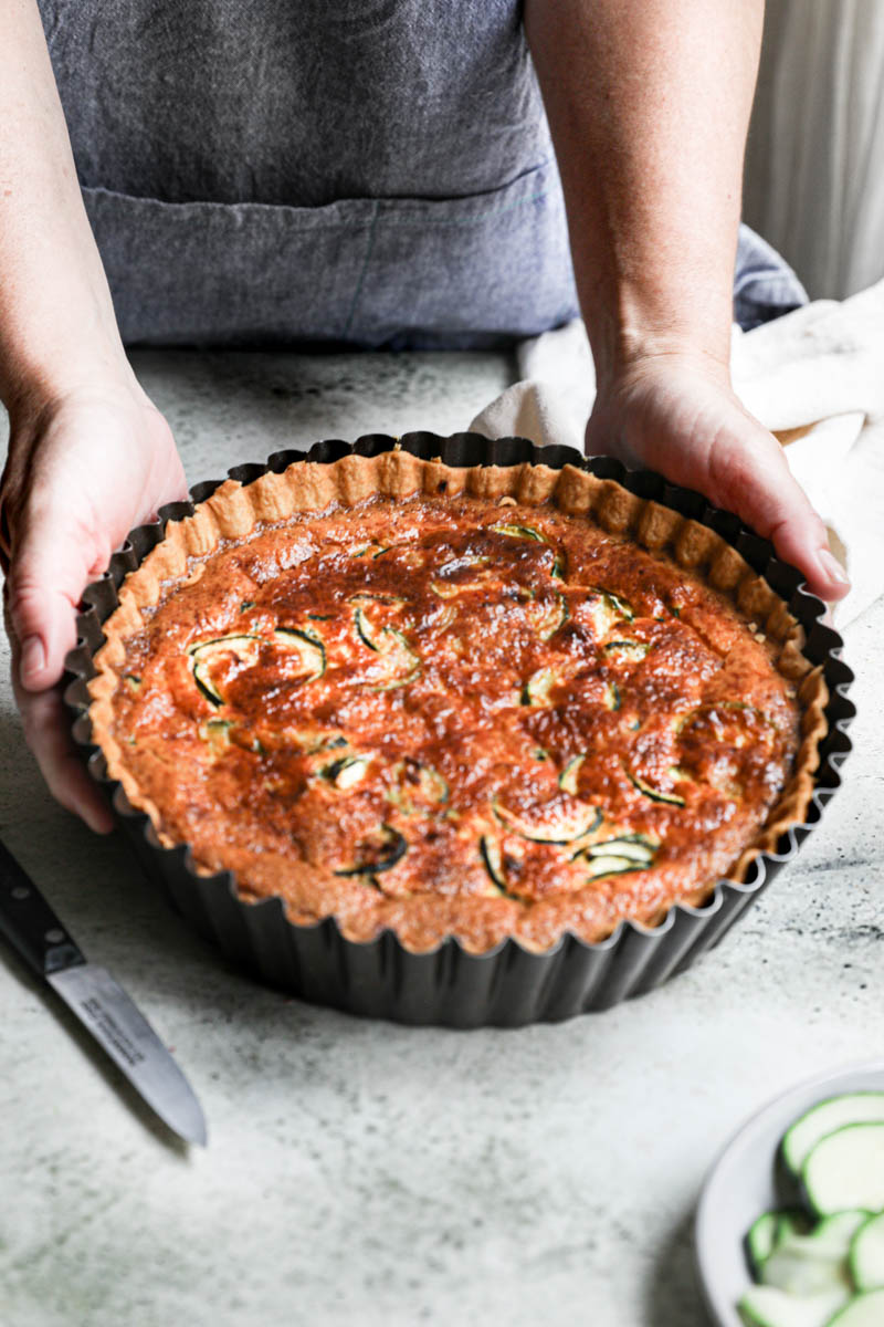 2 hands holding the zucchini quiche inside the tart tin with some sliced zucchinis in the front bottom corner, a black knife on the side and a linen on the back.