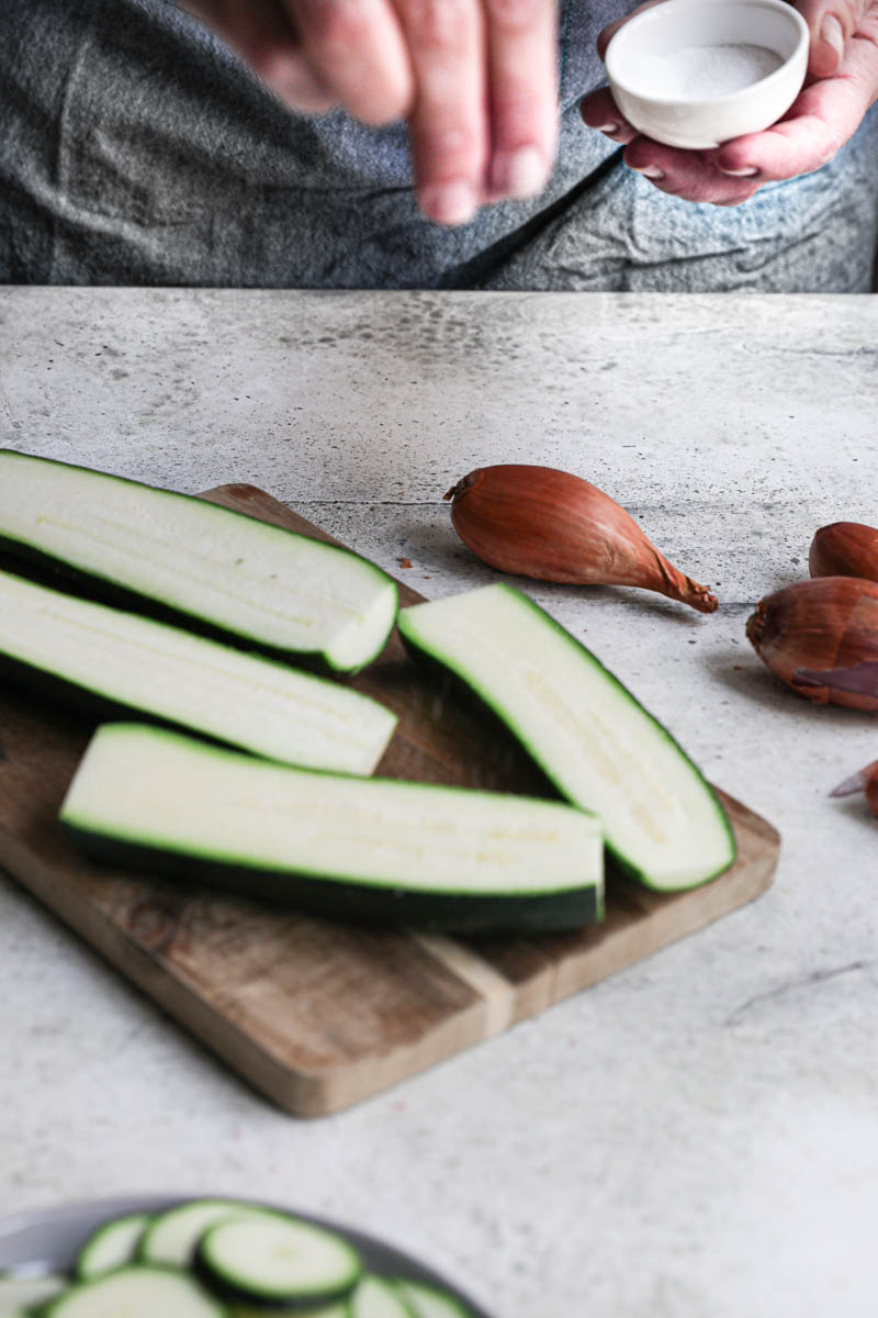 2 hands salting the halved zucchinis placed on a wooden board with some shallots on the side.