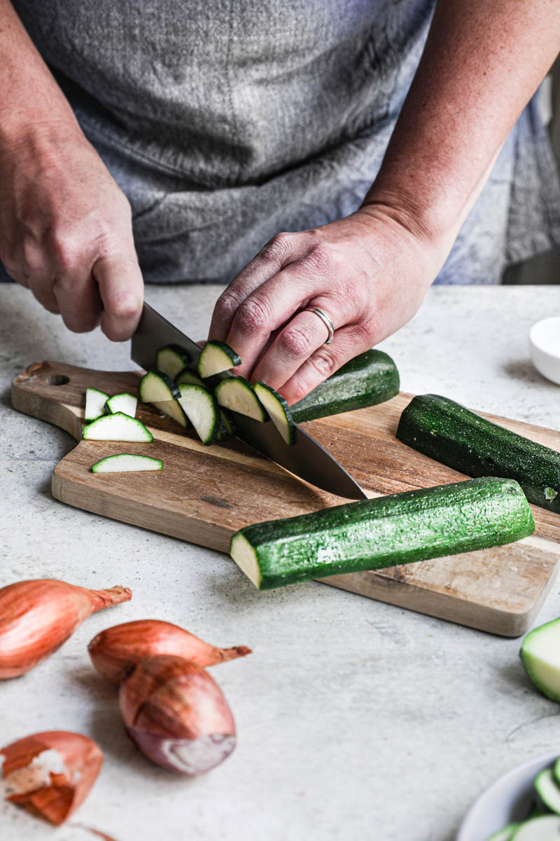 2 hands holding a chef's knife slicing the zucchinis placed on a wooden board with some shallots on the side.