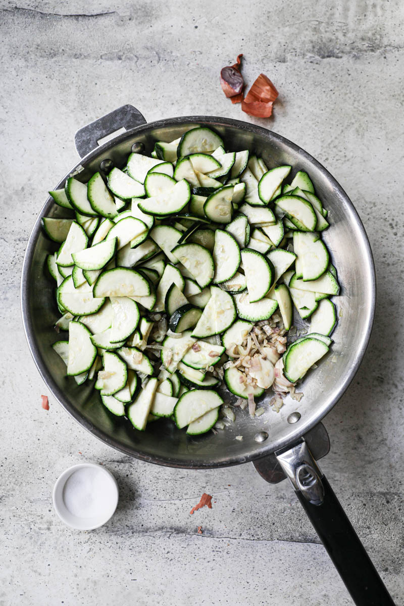 Cooked chopped shallots and sliced raw zucchini on a pan with a dollop of olive oil and small bowl holding salt on the side.